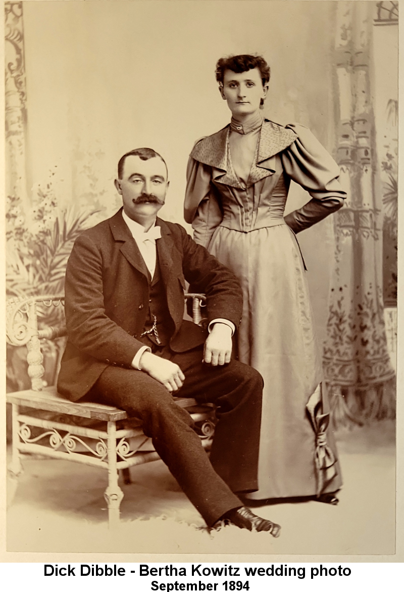 Dick Dibble - Bertha Kowitz wedding photo September 1894; sepia-tone studio photo. Dick Dibble, a slightly insouciant grin on his lips, in dark three-piece suit with white tie, watch chain, short combed dark hair and luxuriant dark handlebar moustace sits on a low table; to his left stands a thin young woman in a medium-toned satin dress with tight-fitting closed bodice and wide patterned collar and large flounced long sleeves; her hair is in a long curl around the top of her head and she has a solemn expression on her thin lips.